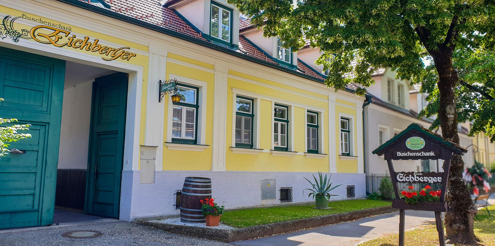 Žltá budova so zelenými dverami a oknami, nápis "Vínny šenky Eichberger", v popredí strom.