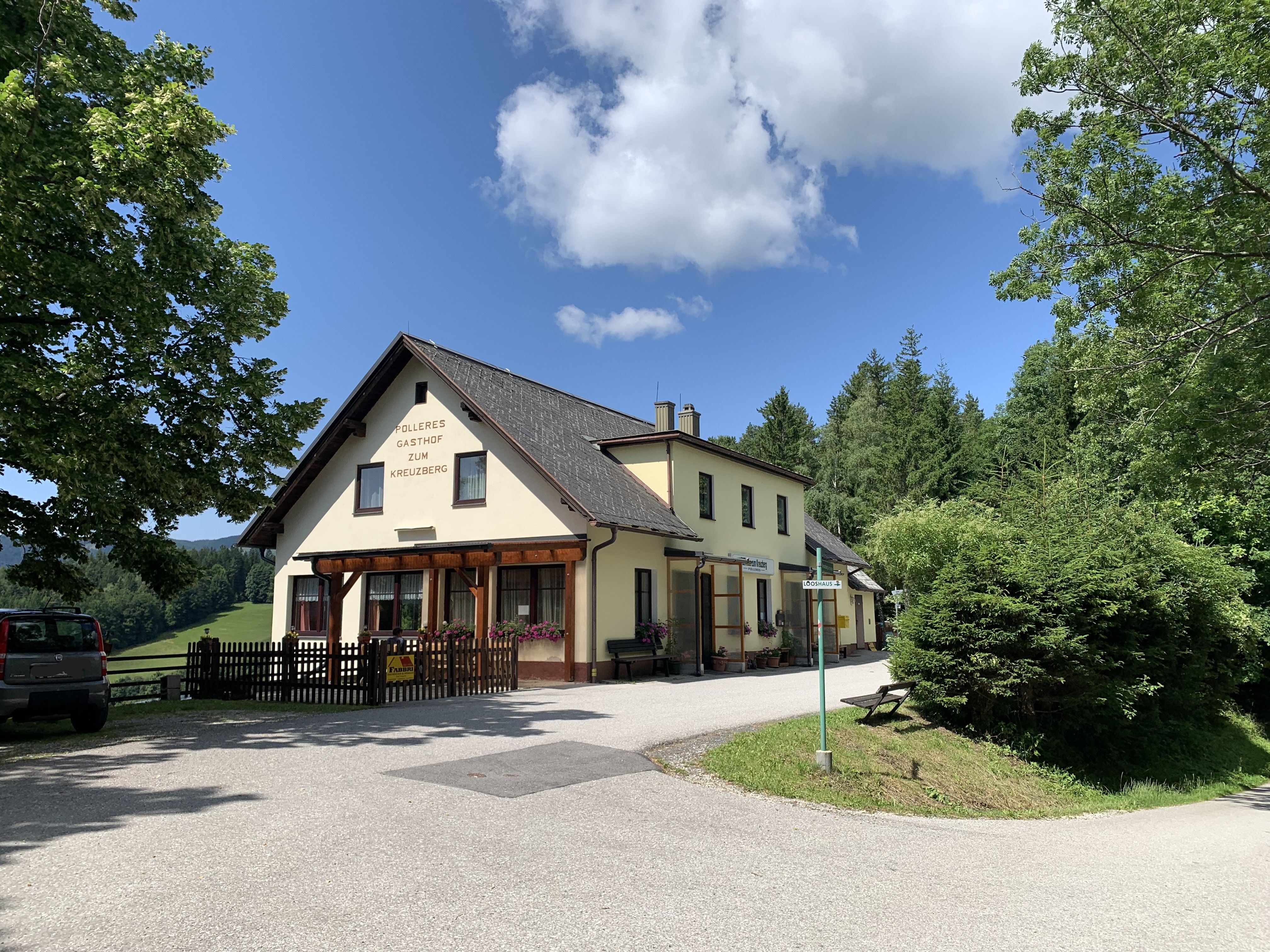 Žltý hostinec s nápisom "Polleres Gasthof zum Kreuzberg" obklopený stromami a modrou oblohou.