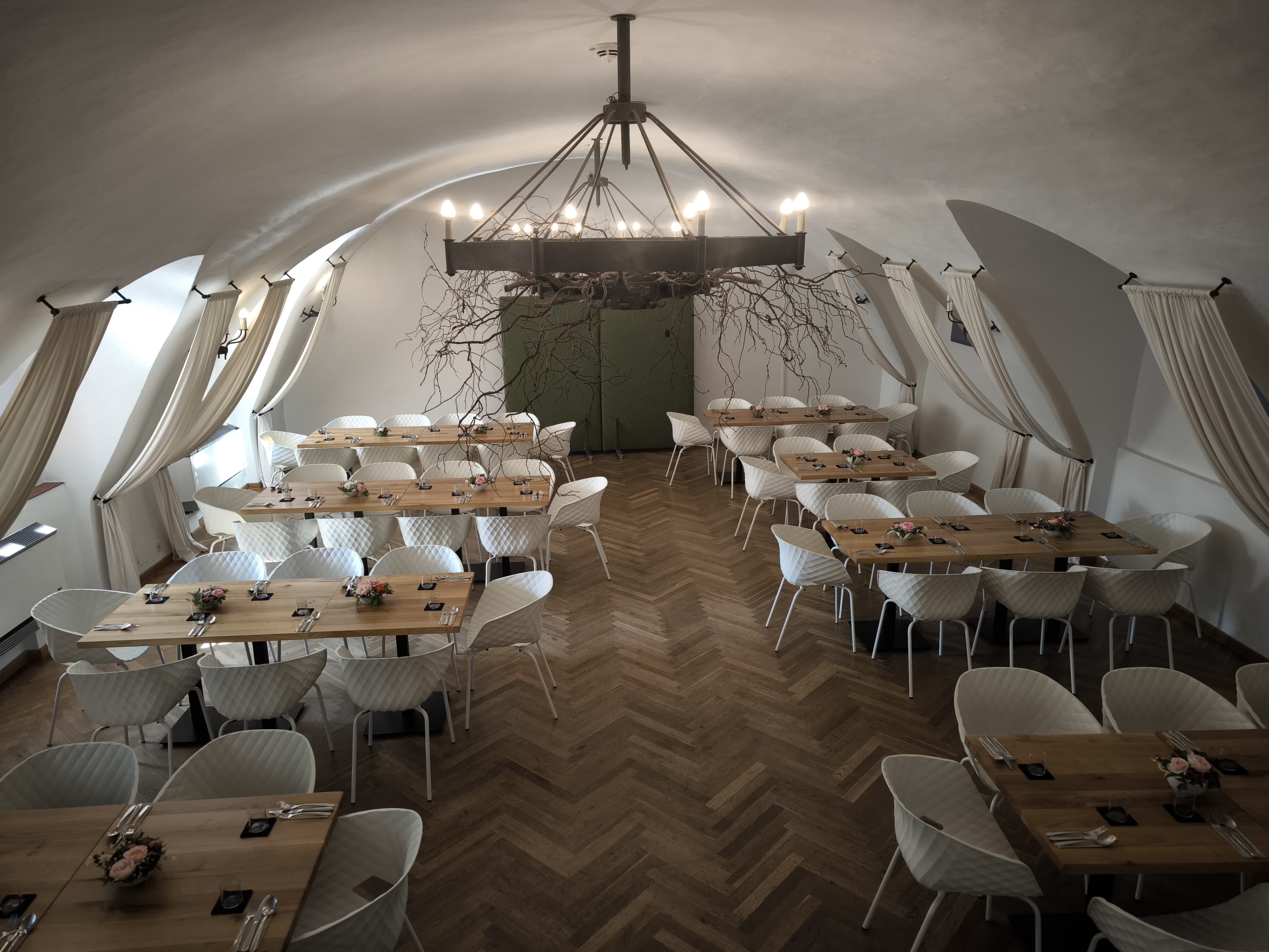 Elegantná sála s drevenými stolmi, parketovou podlahou, bielymi stoličkami a železným lustrom.