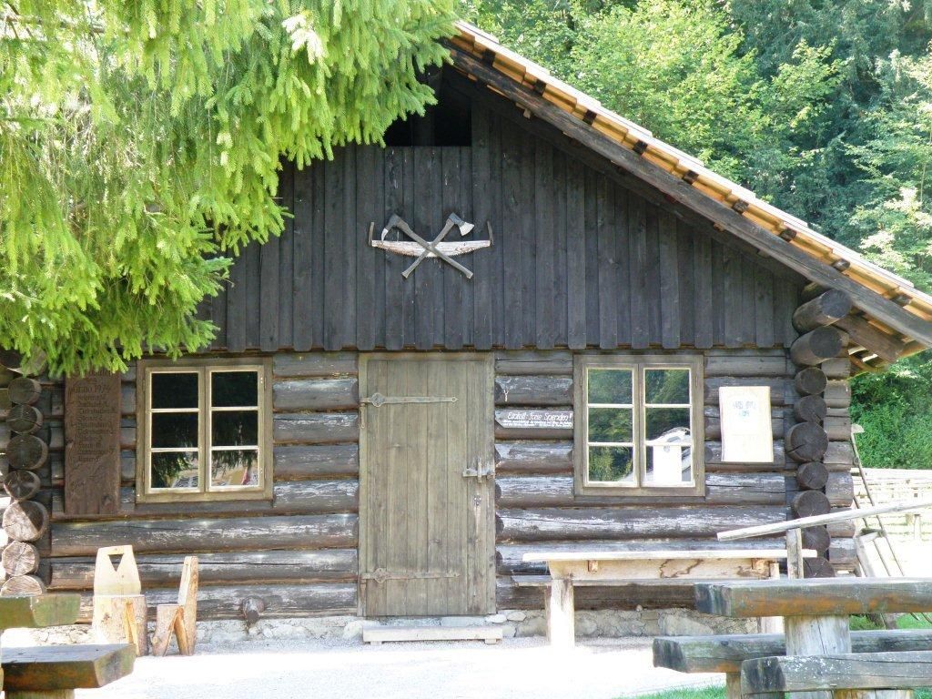 Drevená chatka s dvoma oknami a dverami, obklopená stromami.