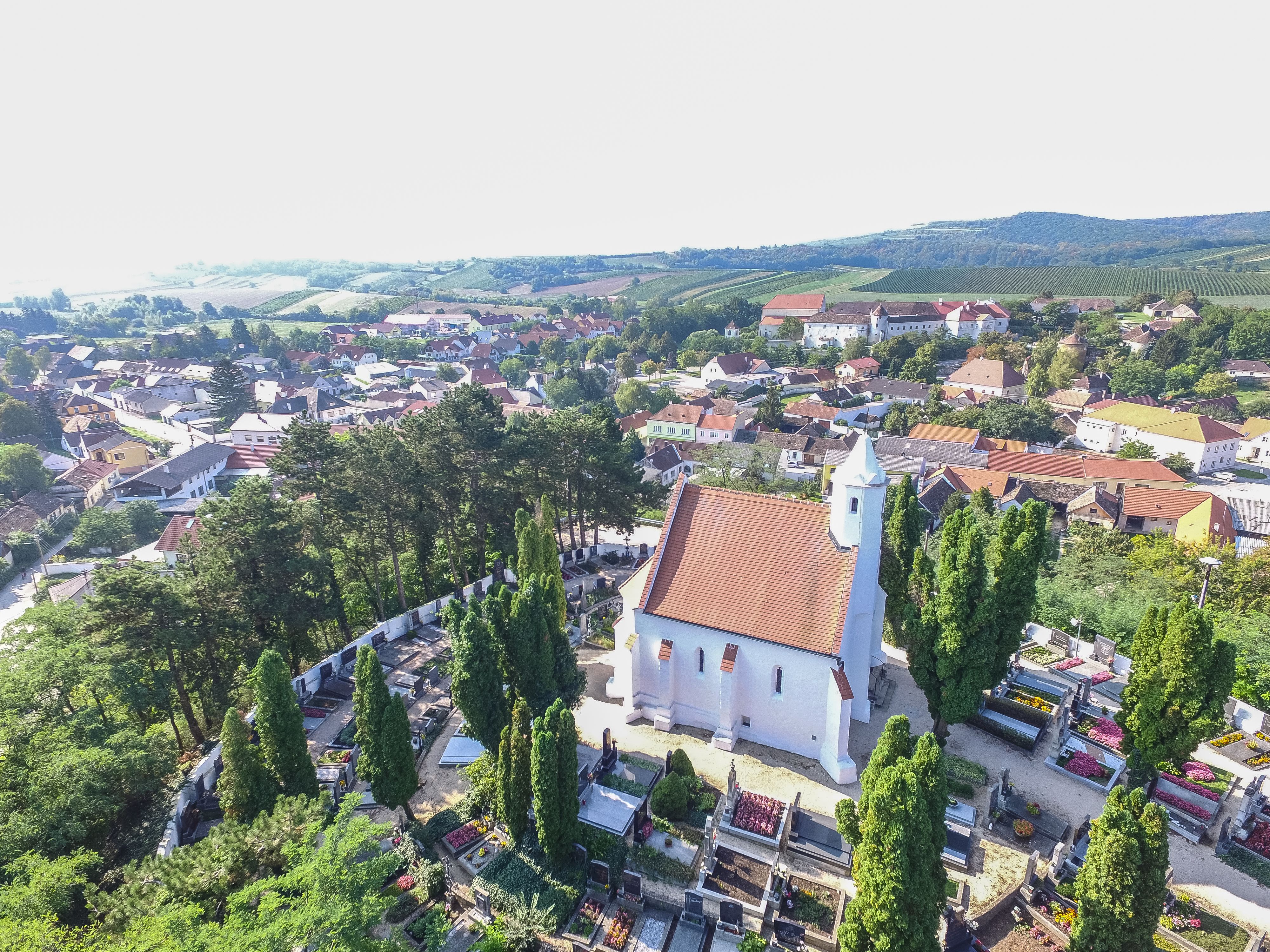 Letecký pohľad na kostol s červenou strechou, obklopený cintorínom a stromami, vo vidieckom prostredí s domami a kopcami v pozadí.