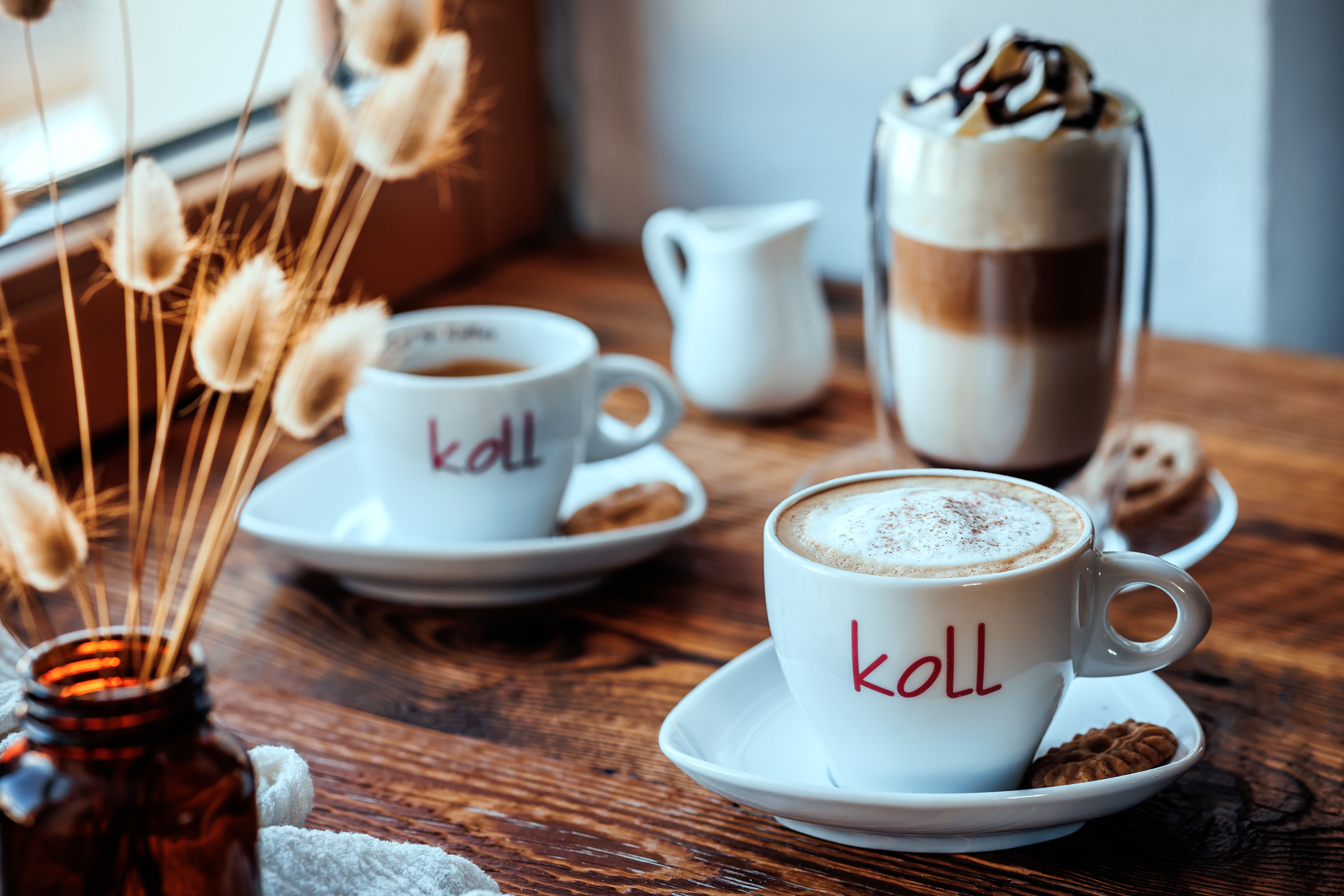 Dve šálky kávy na drevenom stole, jedna s mliečnou penou, pohár latte macchiato a džbán vedľa.