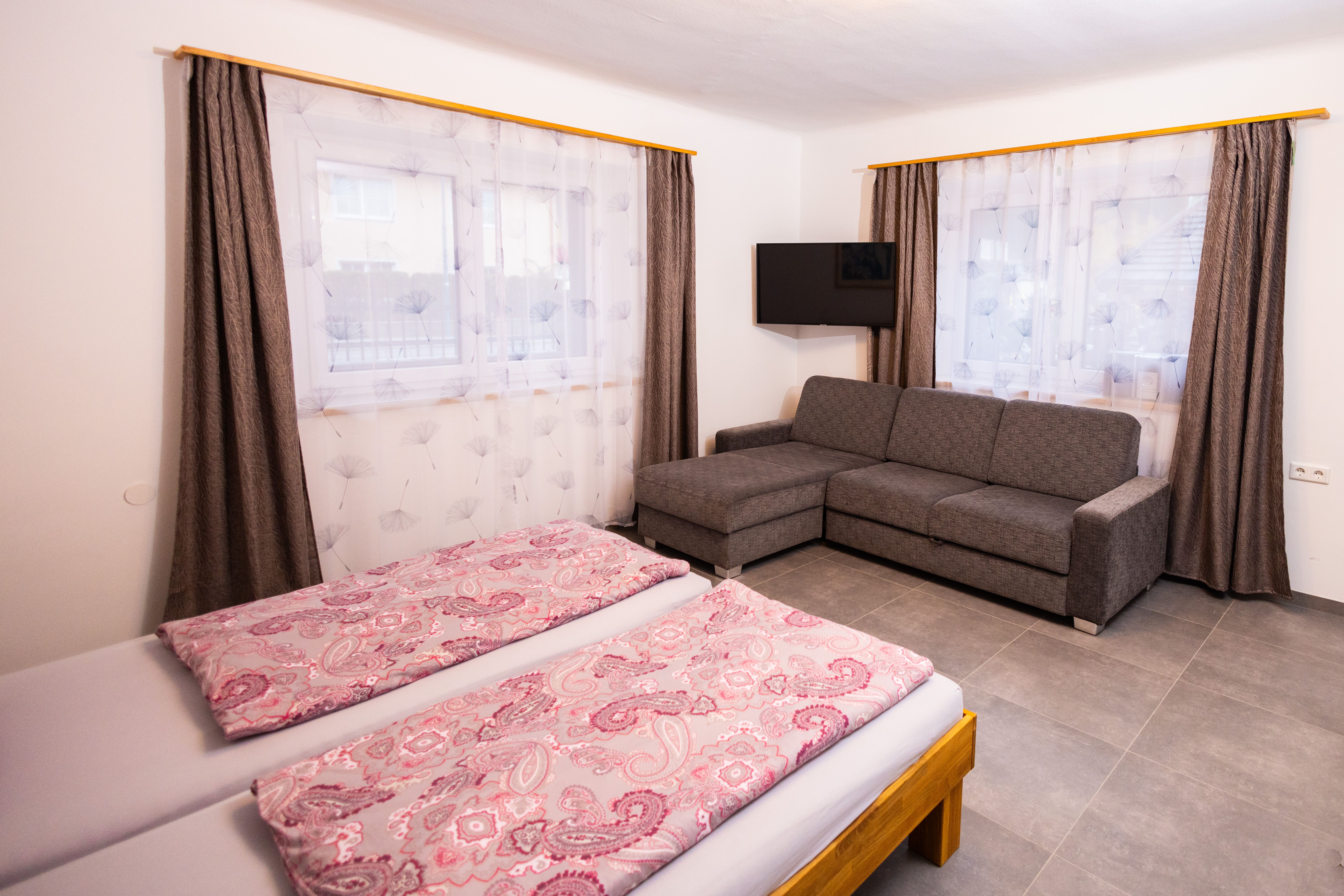 Jedna spálňa s manželskou posteľou, sivou pohovkou, TV a dvoma oknami so závesmi.