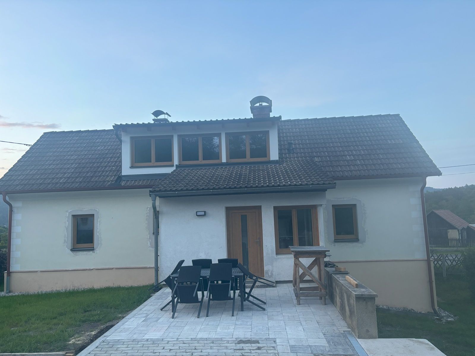 Dvojpodlažný rekreačný dom so šindľovou strechou a terasou, obklopený trávnikom a vidieckym prostredím.