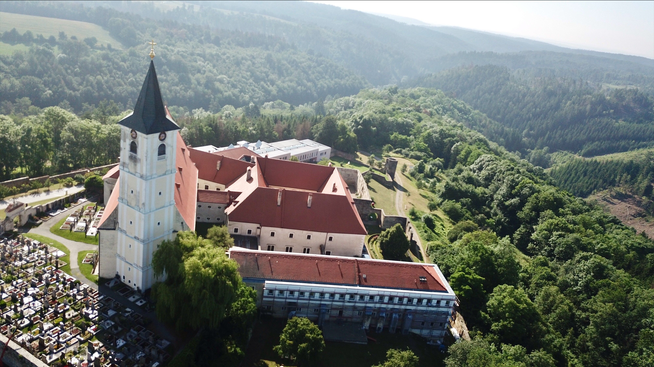 Letecký pohľad na kláštor s červenou strechou a cintorín vedľa neho, obklopený zalesnenými kopcami.