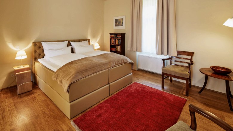 Útulná spálňa s veľkou posteľou, dvoma nočnými stolíkmi s lampami, červeným kobercom a dreveným nábytkom.