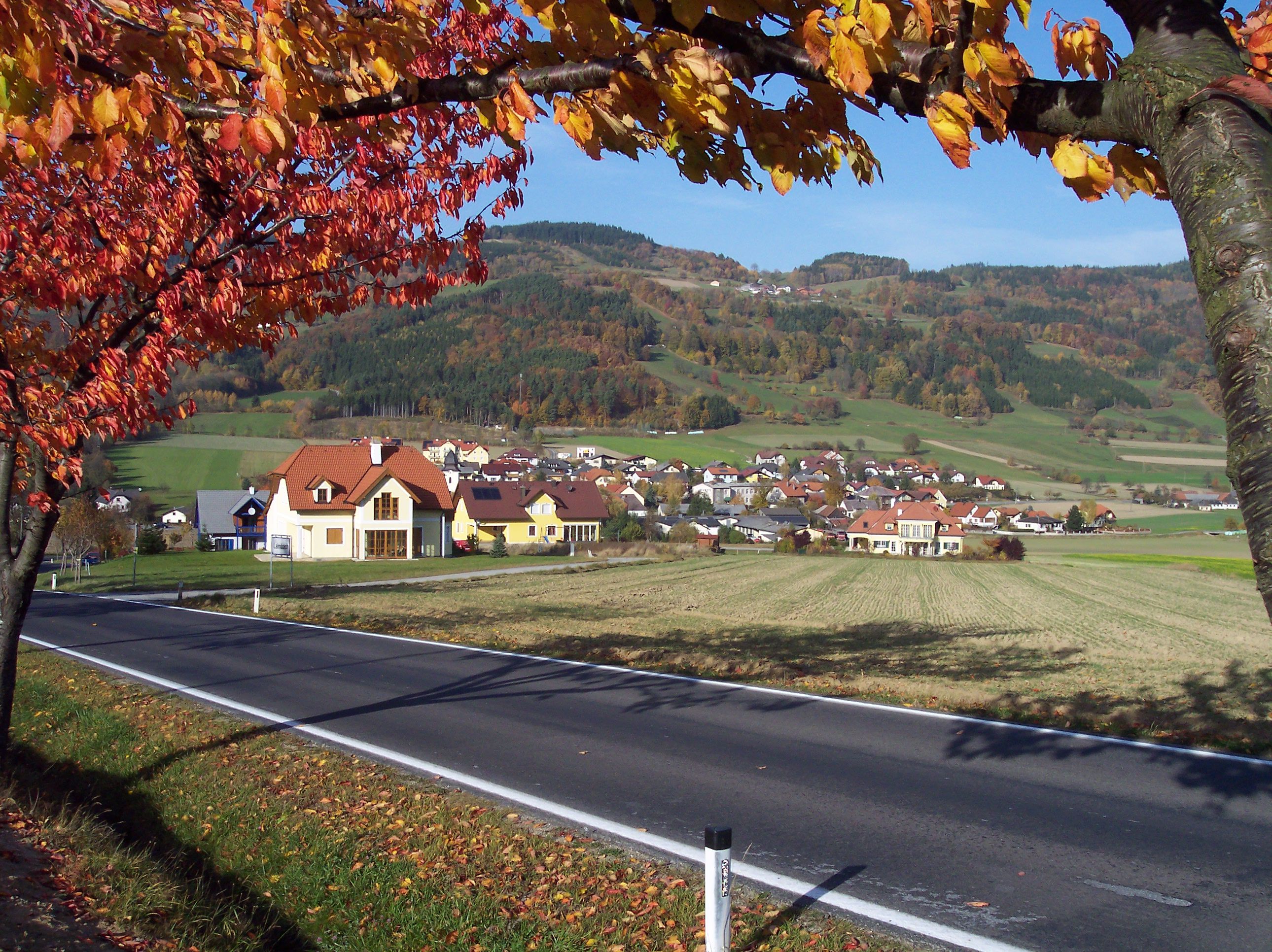 Jesenná krajina s dedinou a kopcami v pozadí, listnaté stromy s farebným lístím v popredí.