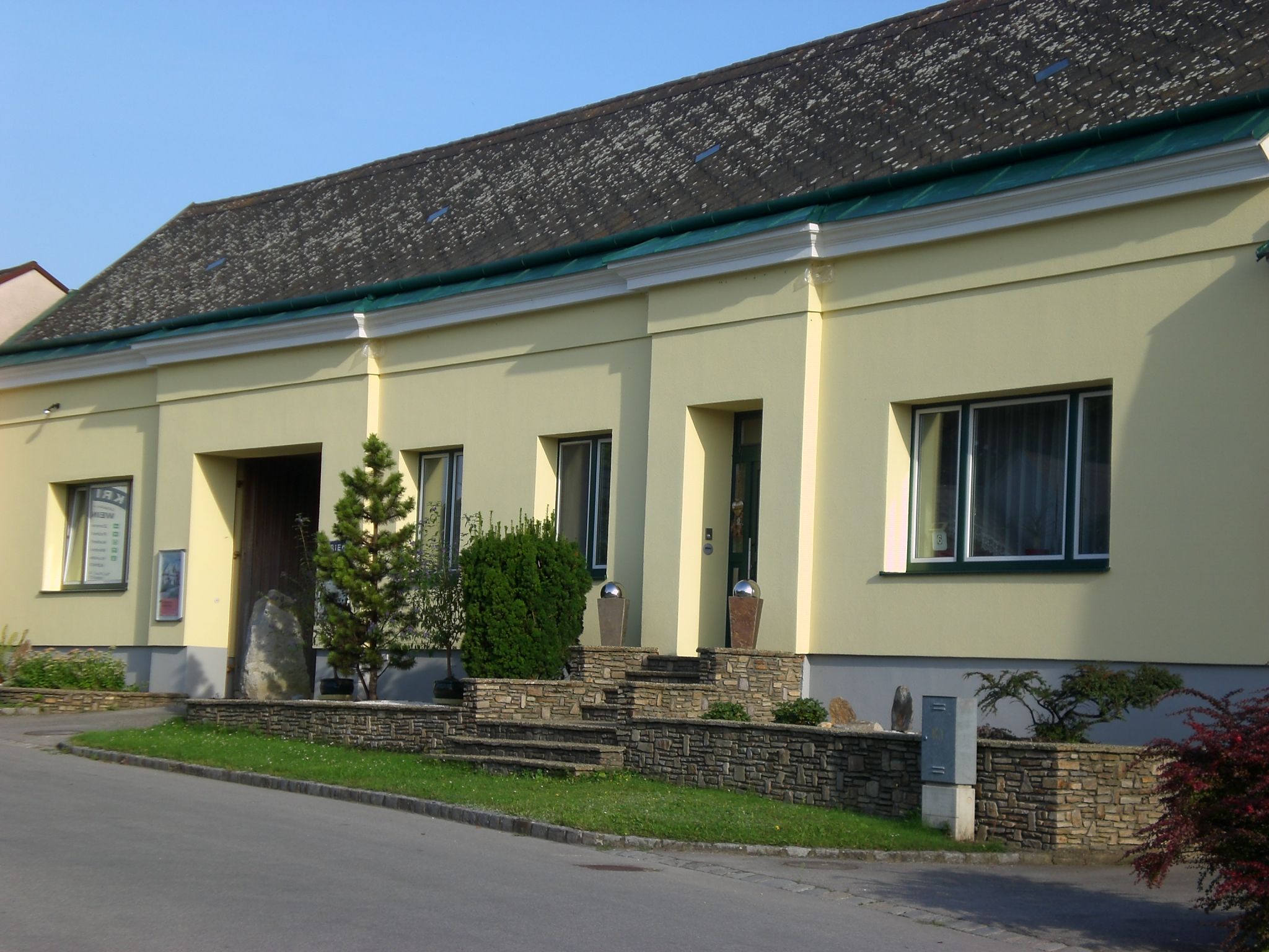 Žltá budova s kamenným múrom a rastlinami pred vchodom.